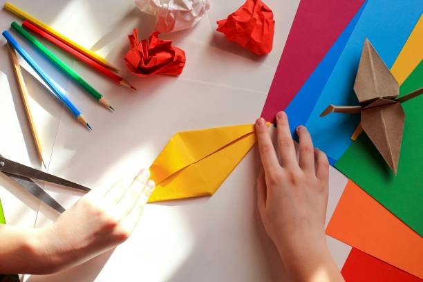 istock Children’s hands doing origami crane from yellow paper on white background with various school supplies. Step-by-step tutorial of origami. Concept of children's creativity, back to school. 1227093802