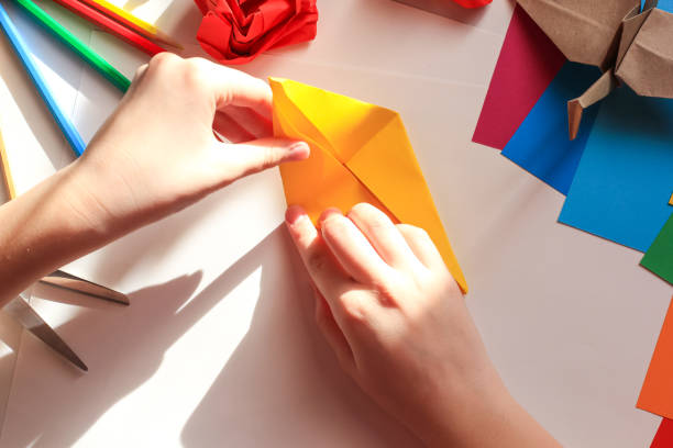 istock Children’s hands doing origami crane from yellow paper on white background with various school supplies. Step-by-step tutorial of origami. Concept of children's creativity, back to school. 1227093815