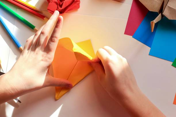 istock Children’s hands doing origami crane from yellow paper on white background with various school supplies. Step-by-step tutorial of origami. Concept of children's creativity, back to school. 1227093817