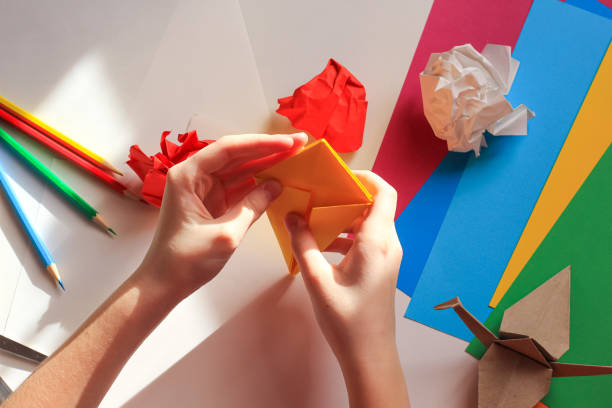 istock Children’s hands doing origami crane from yellow paper on white background with various school supplies. Step-by-step tutorial of origami. Concept of children's creativity, back to school. 1227093820