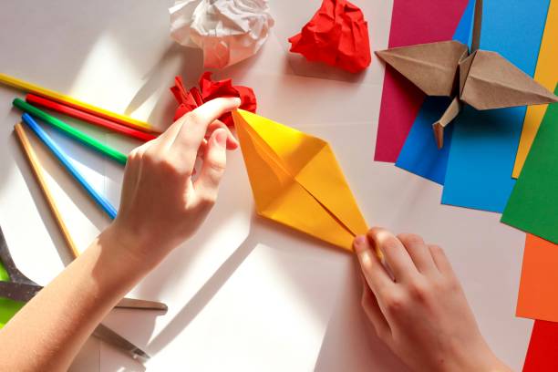 istock Children’s hands doing origami crane from yellow paper on white background with various school supplies. Step-by-step tutorial of origami. Concept of children's creativity, back to school. 1227093833