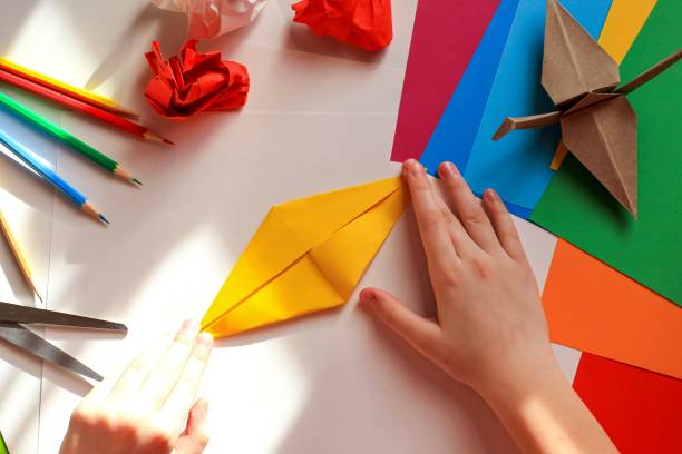 istock Children’s hands doing origami crane from yellow paper on white background with various school supplies. Step-by-step tutorial of origami. Concept of children's creativity, back to school. 1227093836