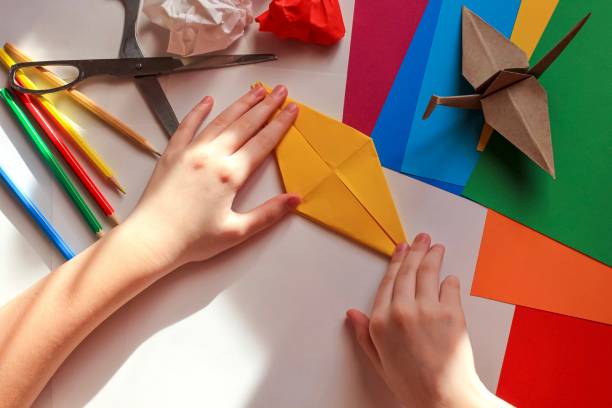 istock Children’s hands doing origami crane from yellow paper on white background with various school supplies. Step-by-step tutorial of origami. Concept of children's creativity, back to school. 1227093844