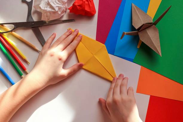 istock Children’s hands doing origami crane from yellow paper on white background with various school supplies. Step-by-step tutorial of origami. Concept of children's creativity, back to school. 1227093847