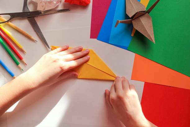 istock Children’s hands doing origami crane from yellow paper on white background with various school supplies. Step-by-step tutorial of origami. Concept of children's creativity, back to school. 1227093855