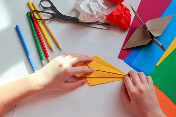 Children’s hands doing origami crane from yellow paper on white background with various school supplies. Step-by-step tutorial of origami. Concept of children's creativity, back to school.