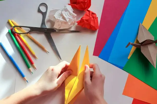 Children’s hands doing origami crane from yellow paper on white background with various school supplies. Step-by-step tutorial of origami. Concept of children's creativity, back to school.