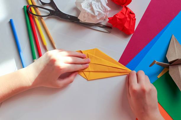 istock Children’s hands doing origami crane from yellow paper on white background with various school supplies. Step-by-step tutorial of origami. Concept of children's creativity, back to school. 1227093881