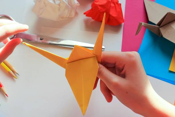 istock Children’s hands doing origami crane from yellow paper on white background with various school supplies. Step-by-step tutorial of origami. Concept of children's creativity, back to school. 1227093898