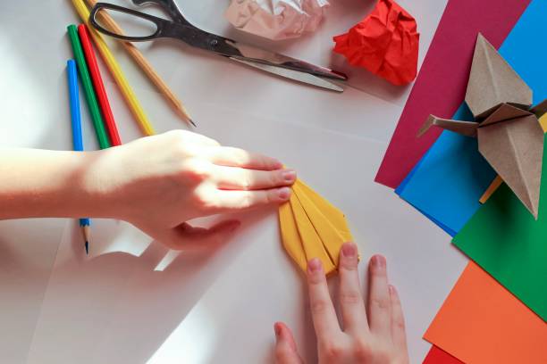 istock Children’s hands doing origami crane from yellow paper on white background with various school supplies. Step-by-step tutorial of origami. Concept of children's creativity, back to school. 1227093900
