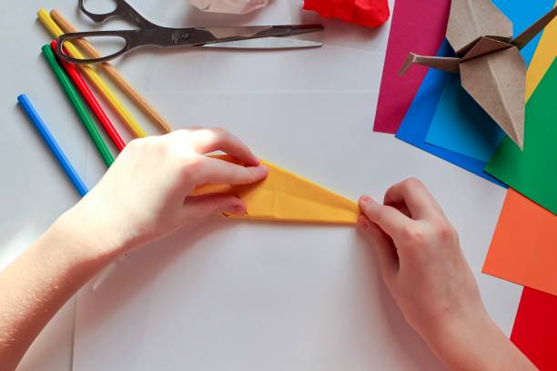 istock Children’s hands doing origami crane from yellow paper on white background with various school supplies. Step-by-step tutorial of origami. Concept of children's creativity, back to school. 1227093909