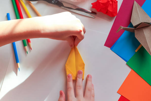 istock Children’s hands doing origami crane from yellow paper on white background with various school supplies. Step-by-step tutorial of origami. Concept of children's creativity, back to school. 1227093911