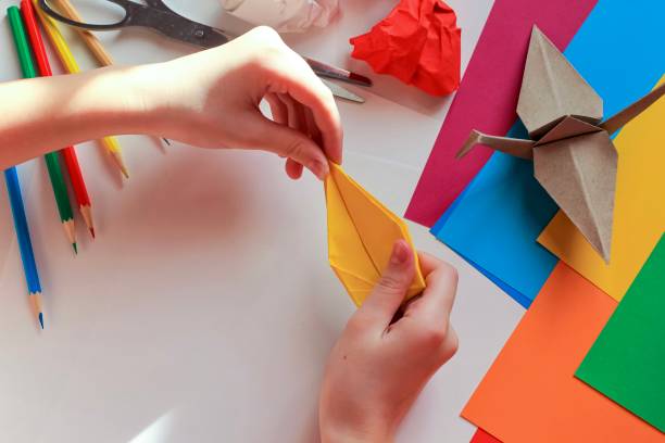 istock Children’s hands doing origami crane from yellow paper on white background with various school supplies. Step-by-step tutorial of origami. Concept of children's creativity, back to school. 1227093925
