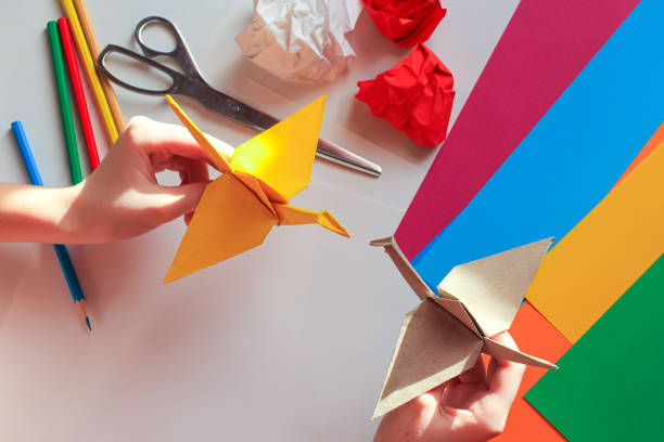 istock Children’s hands doing origami crane from yellow paper on white background with various school supplies. Step-by-step tutorial of origami. Concept of children's creativity, back to school. 1227093936