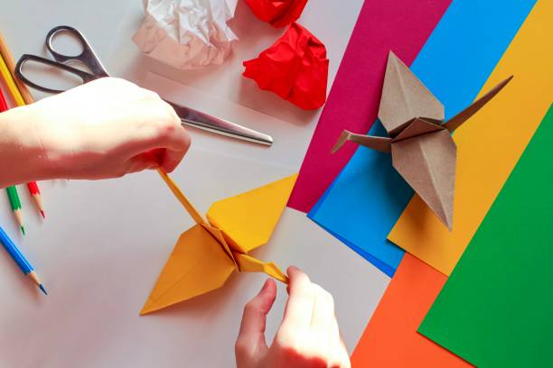 istock Children’s hands doing origami crane from yellow paper on white background with various school supplies. Step-by-step tutorial of origami. Concept of children's creativity, back to school. 1227093949