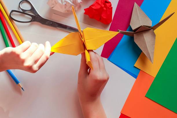 istock Children’s hands doing origami crane from yellow paper on white background with various school supplies. Step-by-step tutorial of origami. Concept of children's creativity, back to school. 1227093956