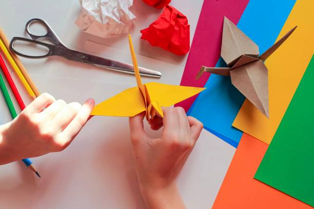 istock Children’s hands doing origami crane from yellow paper on white background with various school supplies. Step-by-step tutorial of origami. Concept of children's creativity, back to school. 1227093974