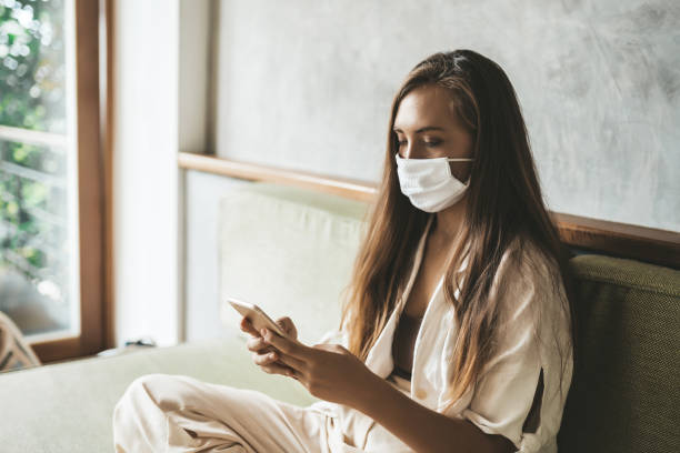 istock Young woman with protective mask using phone sitting on the sofa at home 1227096898