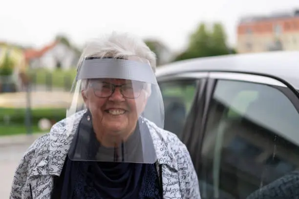 smiling senior woman belonging to vulnerable group of people, with protective face shield besides the car in times of coronavirus beginning to loosen the lockdown