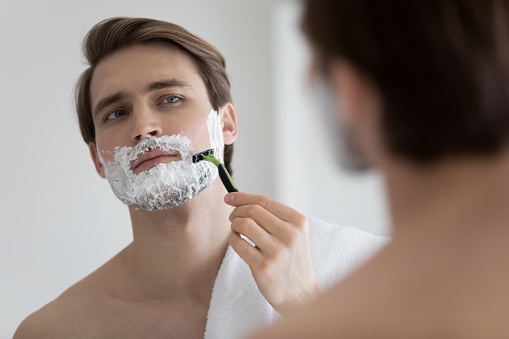 Young caucasian man look in bathroom mirror apply foam gel shaving with razor after shower, millennial male take care of bristle or beard, do morning facial beauty procedure in bath, skincare concept
