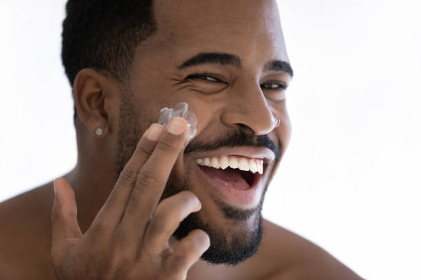 fermez-vous vers le haut de l’homme biracial de sourire employant la crème hydratante de visage - beauté masculine photos et images de collection