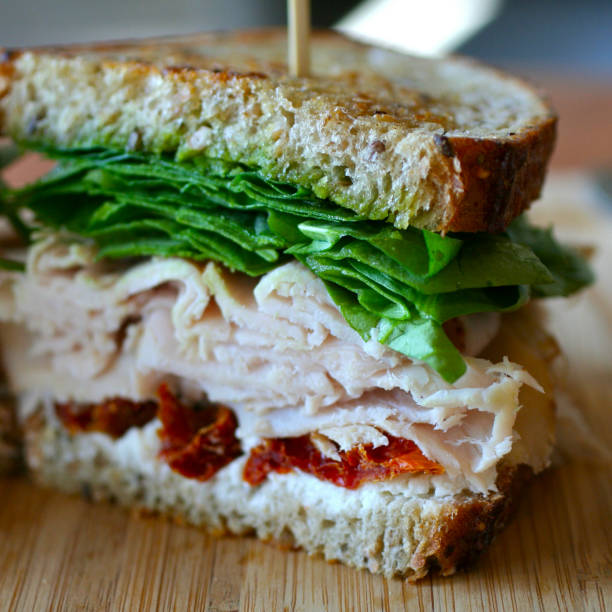 istock Cut sandwich with herbs salad leaves turkey ham and dried tomatoes on a table side view, closeup. Fresh sandwich with ham, cheese, tomatoes and salad leaves ready-to-eat. 1227116880