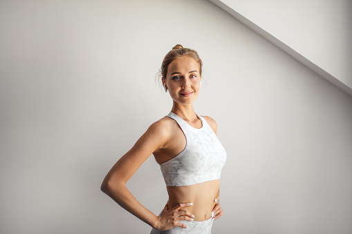 Indoor portrait of young positive sporty female listening to music with wireless earbuds, posing against white wall at Yoga center, taking break after hard workout.