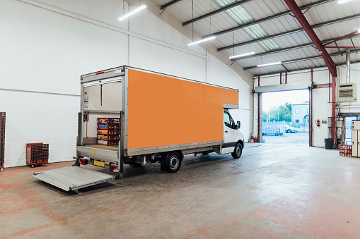 A wide shot of a delivery semi-truck in a distribution warehouse. The back door is open and lowered to the floor, ready for loading.