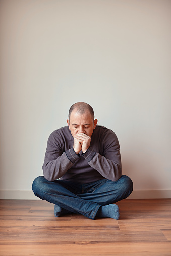 Depressed caucasian man sitting alone on the hardwood floor and leaning against the wall lost in deep thoughts