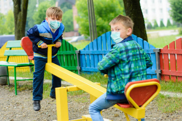 i bambini che studia in maschere mediche giocano in un parco giochi di quarantena durante una pandemia di coronavirus - bench park bench white isolated foto e immagini stock