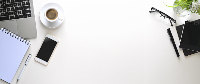 Top view image of white space that surrounded by ceramic coffee cup, black screen smartphone, note, pen, computer laptop, eye glasses, notebook and flowers in glass vase.