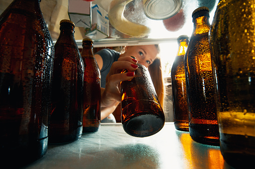 Caucasian woman takes cold refreshing beer from out the fridge, inside view from fridge of hand holding the bottle. Alcohole, domestic life, entertainment, drink concept. Cold cola or beer bottles.