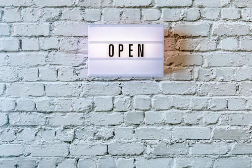 Open sign hanging on wall. Signboard OPEN of lightbox on white vintage brick wall background. Opening sign of cafe, bar, or restaurant. Business open and welcome. Concept of quarantine mitigation