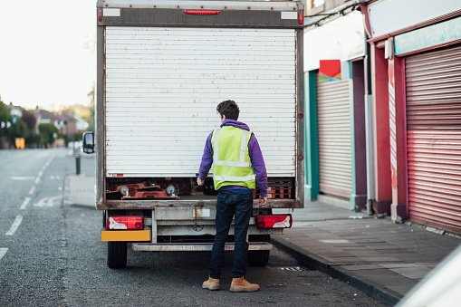 A rear view of a mid adult Caucasian delivery man pulling down the back door on a lorry after making a delivery during the COVID-19 pandemic.