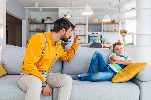 Do not be so naughty. Annoyed man is screaming at his upset son while sitting on sofa at home during the day.