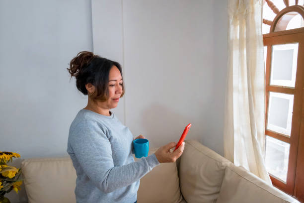 una mujer madura de pie con una taza de café en la sala de estar usando un teléfono celular - using laptop contemplation accessibility contemporary fotografías e imágenes de stock