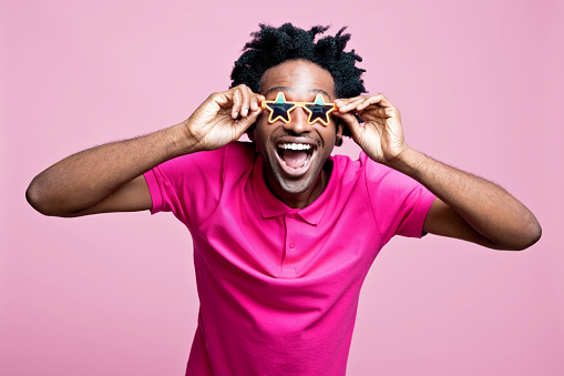 Portrait of happy afro american young man wearing pink polo shirt and star shaped sunglasses, looking at camera. Studio shot on pink background.