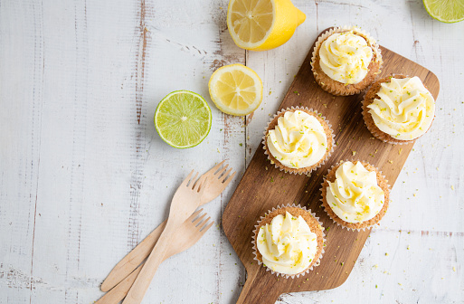 Lemon and poppy seed cupcakes with cheese cream frosting and lemon and lime zest on a rustic white wooden table. Top view