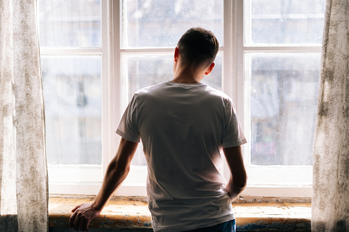 Unrecognizable young man stands by the window on sunny day, enjoying beautiful day, back of view.
