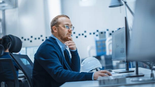 Shot of Male Industrial Engineer Solving Problems, Working on a Personal Computer, He is Working in Office in Modern Factory with High-Tech Machinery.