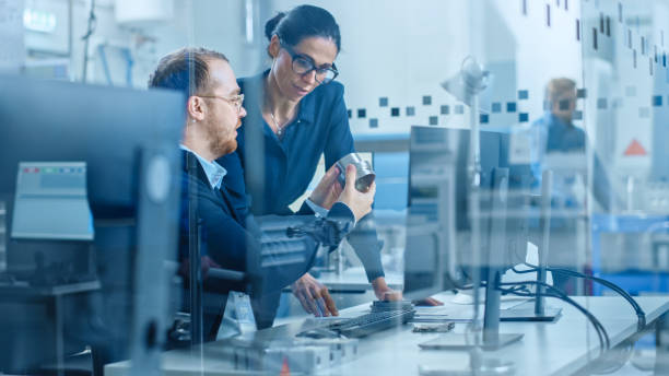 istock Modern Factory: Female Project Supervisor Talks to a Male Industrial Engineer who Works on Computer. They Inspect Machinery Mechanism and Design Improved Version. Working High-Tech Industrial Facility 1227179656