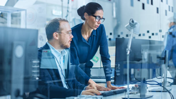 istock Modern Electronics Factory: Female Supervisor Talks to a Male Electrical Engineer who Works on Computer with CAD Software. Developing PCB, Microchips, Semiconductors and Telecommunications Equipment 1227179688