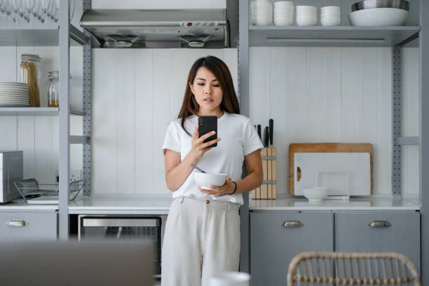istock Young Asian woman using smartphone while enjoying breakfast in the kitchen at home 1227180640