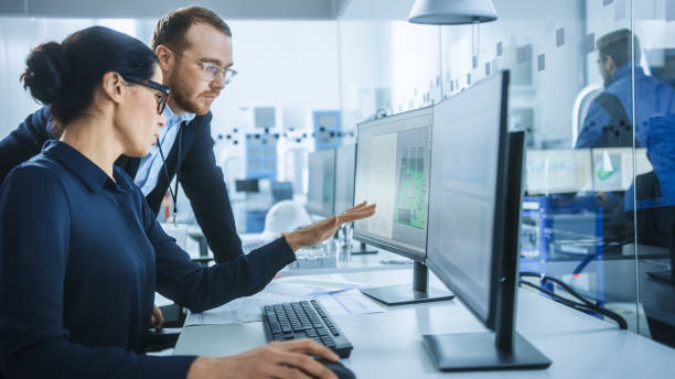 istock Modern Electronics Factory: Male Supervisor Talks to a Female Electrical Engineer who Works on Computer with CAD Software. Developing PCB, Microchips, Semiconductors and Telecommunications Equipment 1227182079