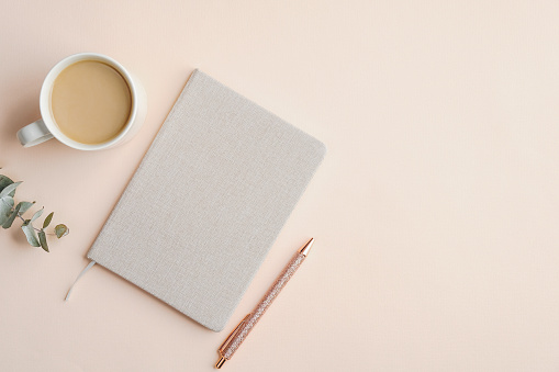 Flat lay, top view office table desk. Feminine workspace with paper notebook, pen, cup of coffee and eucalyptus leaf on beige background with copy space. Business concept