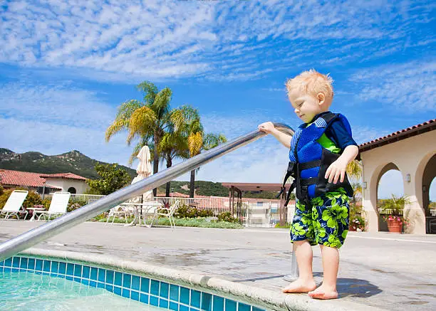 A cute little boy with a life jacket bravely steps into an outdoor swimming pool in San Diego, California