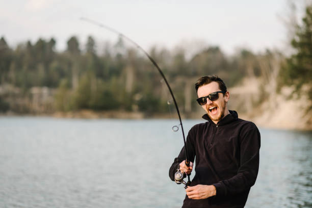 istock Fisherman with rod, spinning reel on the river bank. Man catching fish, pulling rod while fishing from lake or pond with text space at weekend. Fishing for pike, perch, carp on beach lake or pond. 1227191400