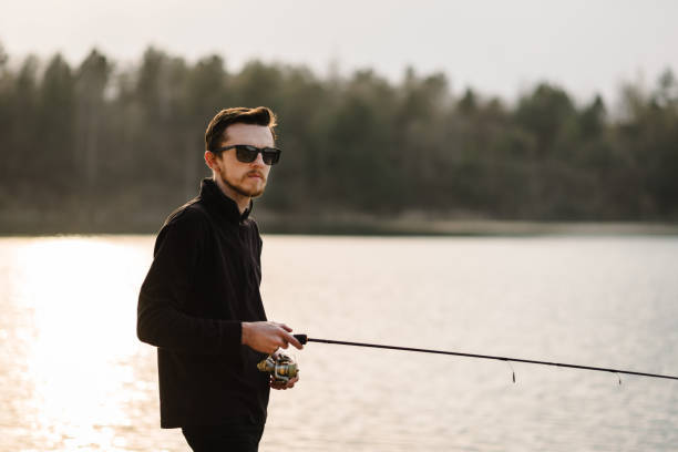 istock Fisherman with rod, spinning reel on the river bank. Man catching fish, pulling rod while fishing from lake or pond with text space at weekend. Fishing for pike, perch, carp on beach lake or pond. 1227191404