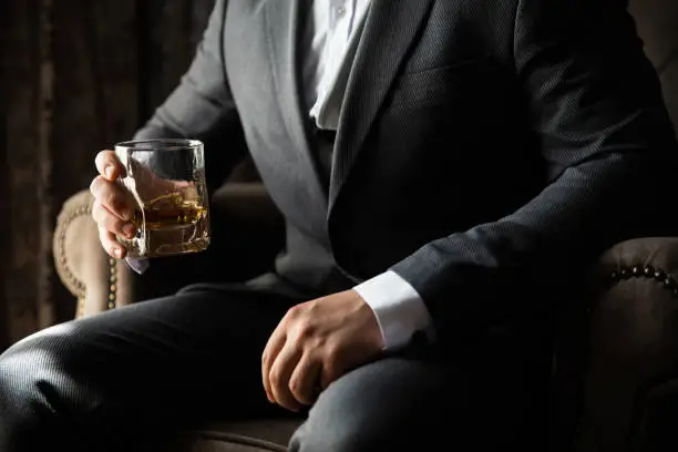 A man in a suit sitting on a chair. In his hands a glass filled with whiskey.