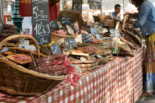 St. Tropez, France - June 2019: Variety of delicious salami in baskets with price tags at the traditional street food market. Open air sales booth or market stall, people buying salami.